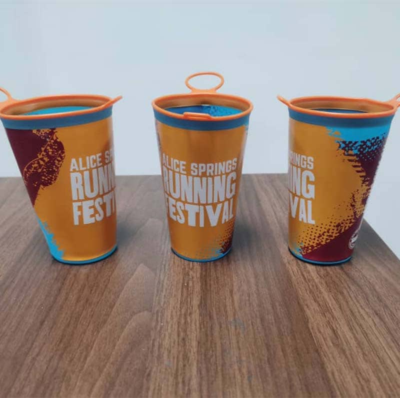 Three reusable cups with orange lids, featuring the "Alice Springs Running Festival" text in white, stand on a wooden surface against a plain light background—perfect custom merchandise or promotional products for your next event.